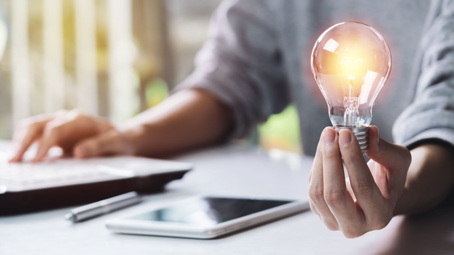 A person holding a light bulb with tablet and laptop on desk.