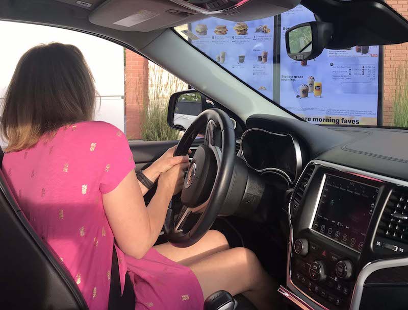 Person sitting in care ordering from an outdoor digital menu board.