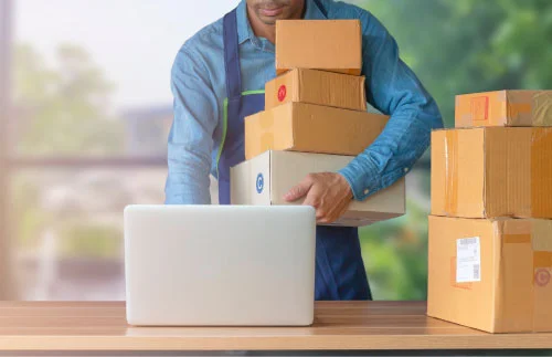 A person holding 4 boxes with more boxes stacked next to a laptop.